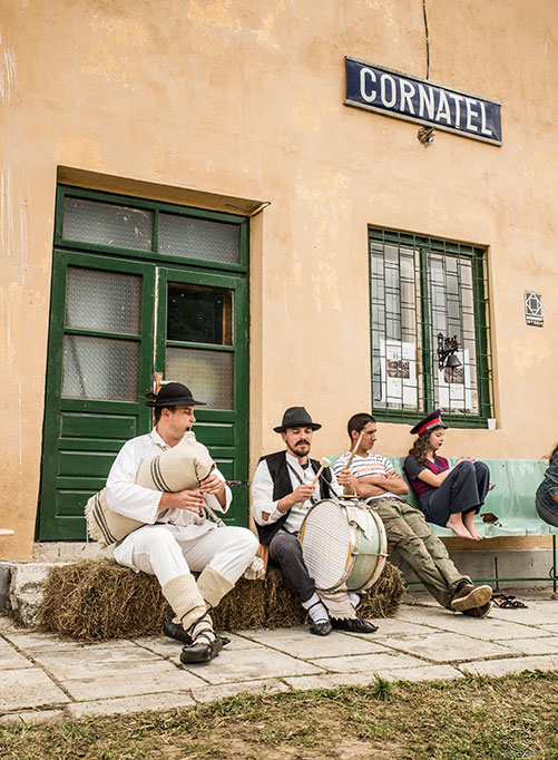 Ein Dudelsackspieler und ein Trommler in Folklorekleidung spielen vor dem Bahnhofsgebäude von Cornatel