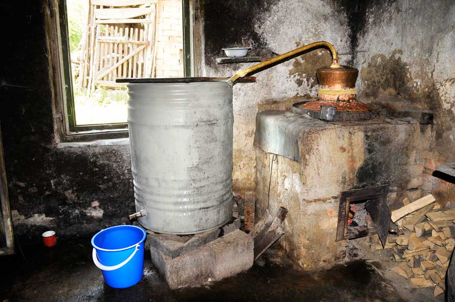 großes Blechfass steht erhöht auf zwei Steinen und ist über eine Kupferleitung mit einem danebenstehenden Ofen verbunden und unter dem Fass steht ein blauer Plasteeimer