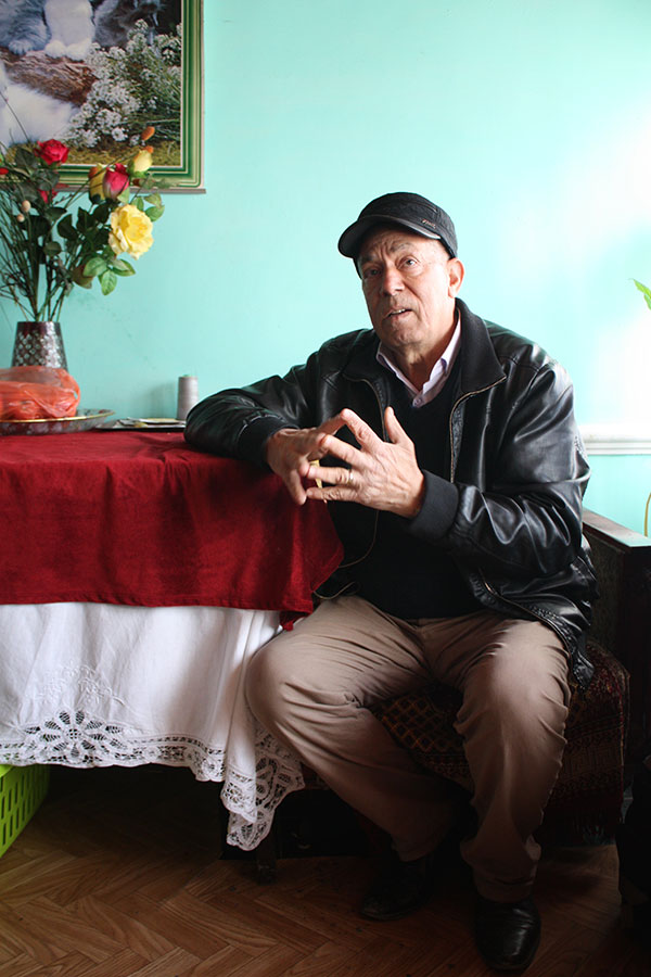 Mann sitzt erzählend an einem Tisch mit roter Decke und einem Blumenstrauß darauf vor einer hellblauen Wand