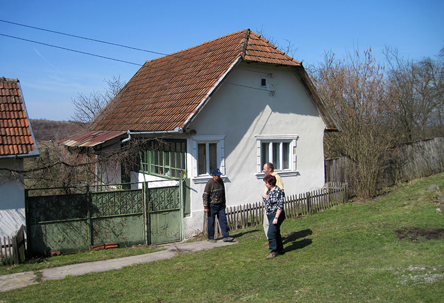 Hella und ihr Mann stehen vor einem Haus und reden mit einem Dorfbewohner