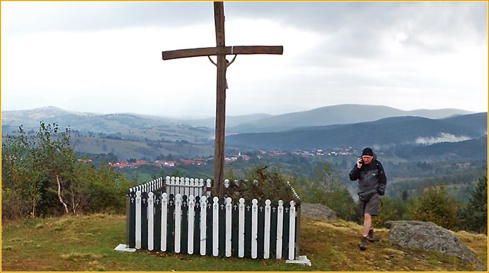 Hans telefoniert neben einem eingezäunten Kreuz auf einem Berg mit Berglandschaft im Hintergrund