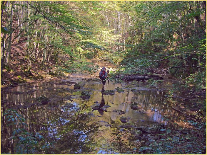 Hans überquert mit großem Wanderrucksack einen Fluss auf Steinen