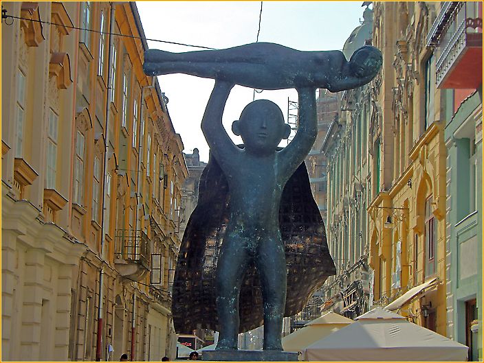 nackter Mann mit offenem Umhang als Bronzefigur hebt eine Frau über seinem Kopf in einer Fußgängerzone