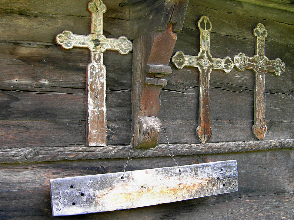 Holzkreuze an einer Holzwand und darunter hängend ein Holzbrett