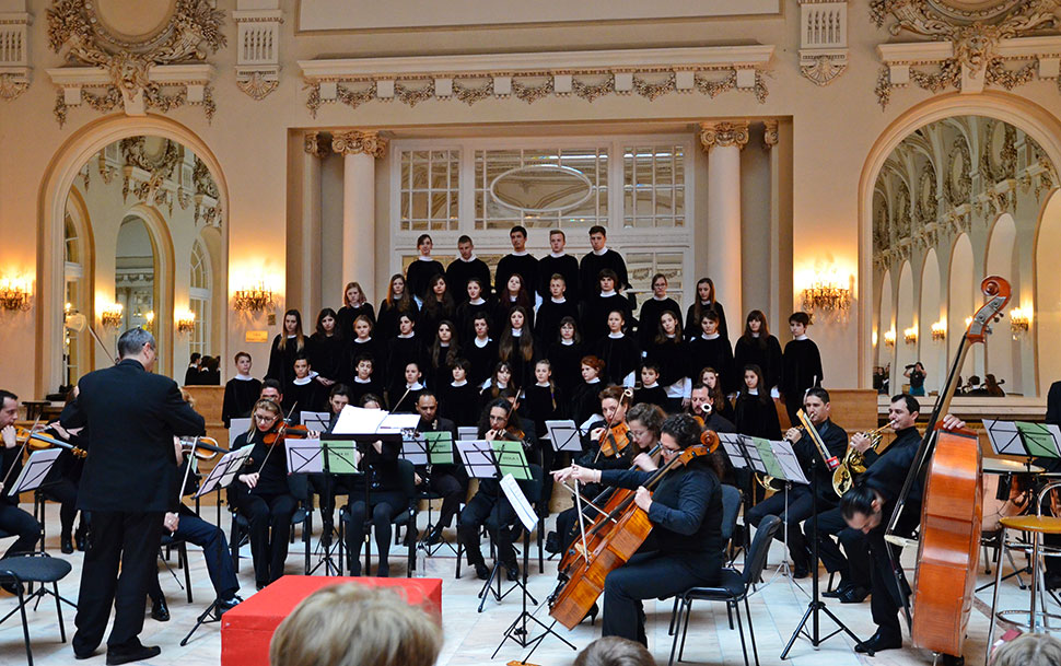 Kinder stehen in schwarzen Umhängen hinter dem Orchester auf der Bühne in einem Barocksaal