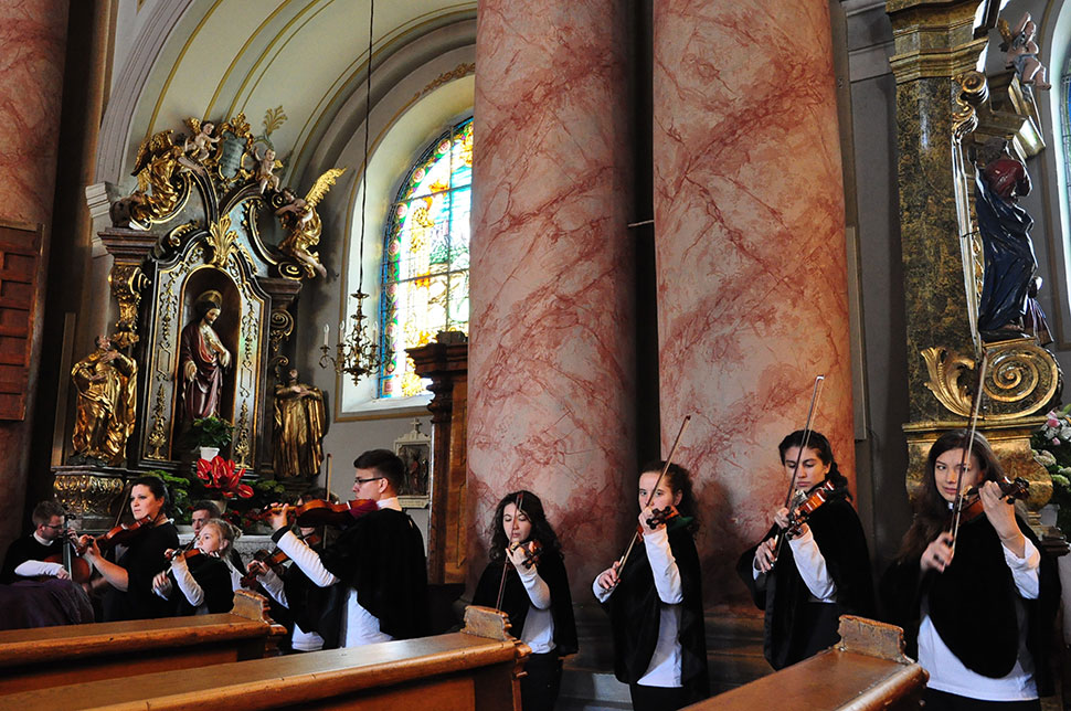 Kinder spielen in schwarzen Umhängen vor Säulen und einem Altar im Hintergrund Violine