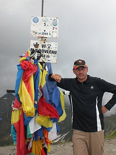 Wanderer lehnt am Gipfelkreuz welches mit rumänischen Flaggen geschmückt ist.