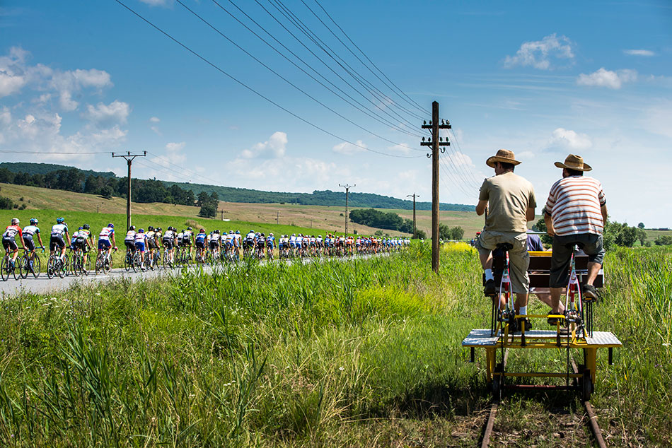Zwei Touristen fahren auf einer Draisine während auf der Straße neben dem Gleis eine Radrenngruppe vorbei fährt