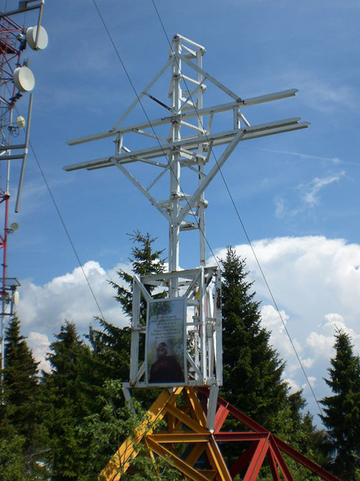 weißes Metallkreuz vor Bäumen und blauen Himmel mit einigen Wolken