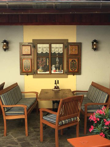 Außenbereich des Restaurant mit Gartenbänken und Stühlen und einem an die Wand gemalten rustikalen Fenster in der Bildmitte