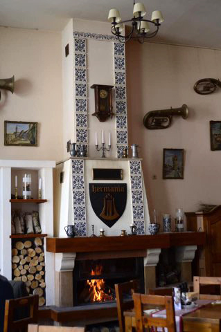 Kamin im Restaurant Hermania mit Feuer, bemalten blauen Keramikfliesen und einer Wanduhr sowie Bildern und Musikinstrumenten an den Wänden