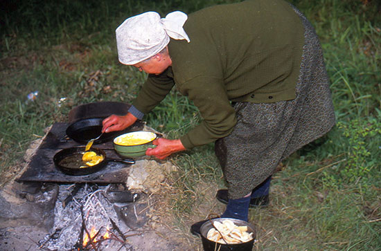 Foto von Feuerstelle mit Pfannen  und einer Frau, welche etwas in die Pfanne legt